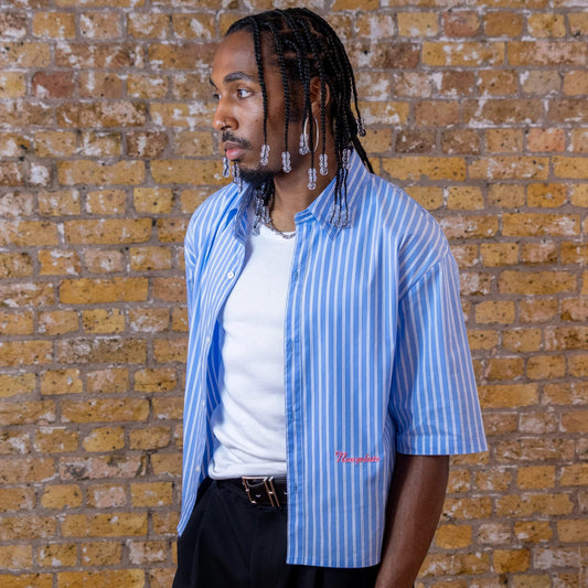 Man wearing a blue striped shirt and black pants standing against a brick wall.